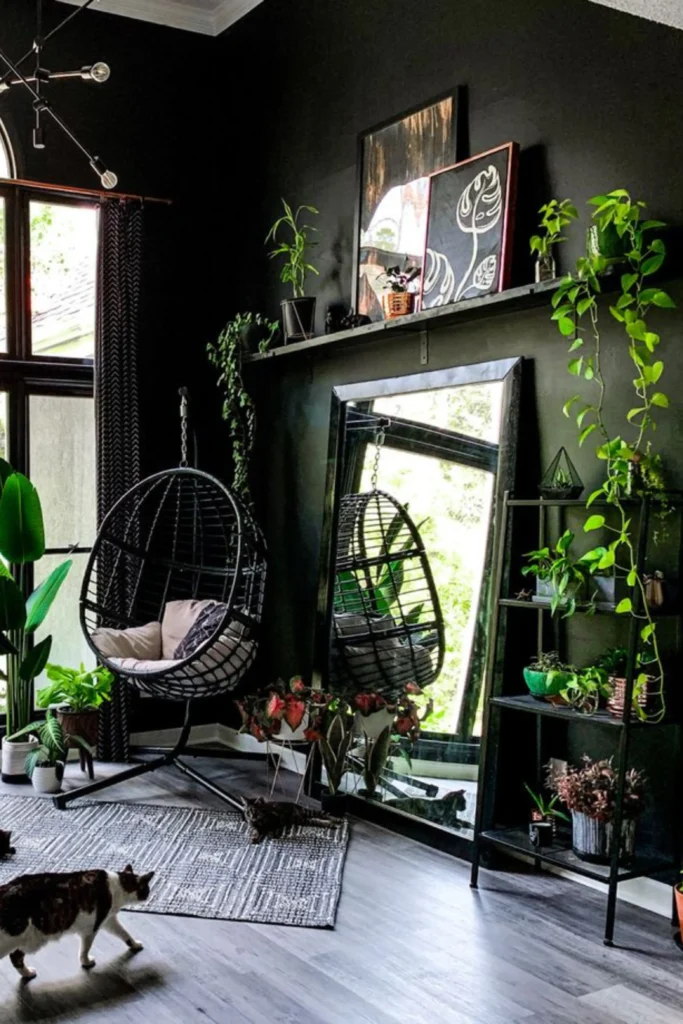 Indoor Botanical Greenery display in a moody room with dark green walls and grey flooring. The setup features various potted plants, including a trailing pothos plant cascading down from a black tiered shelf unit, bringing an instant splash of vibrant color. The space is styled with a black hanging egg chair with cream cushions, a large leaning floor mirror reflecting the room, a textured geometric area rug, and a high vertical shelf displaying botanical framed art and small planters.