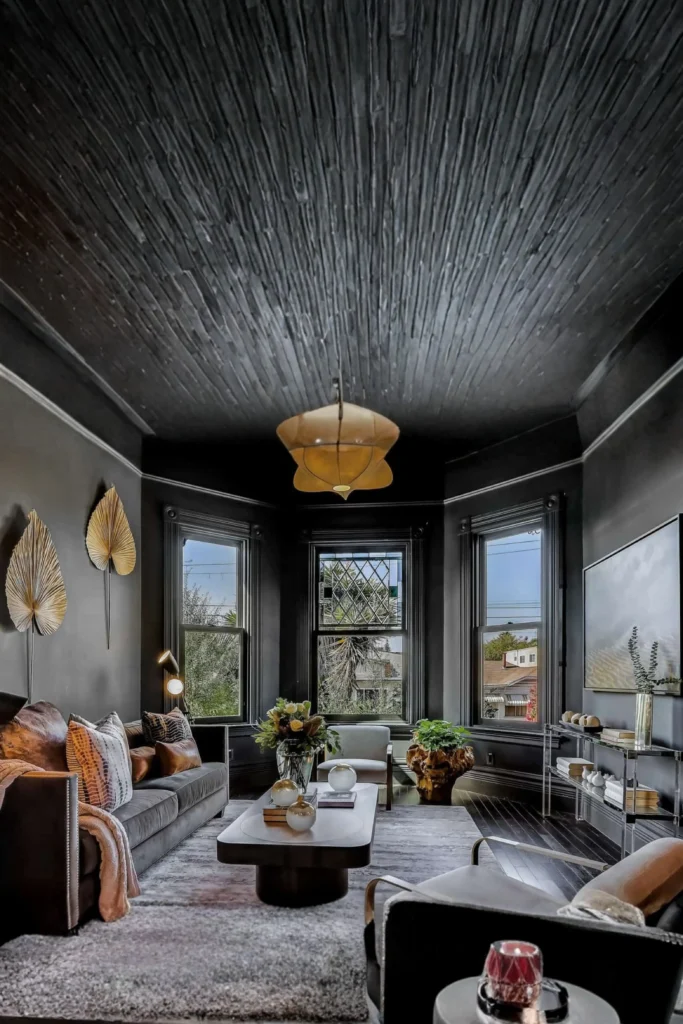 A dramatic, moody living room showcasing how playing with unexpected proportions adds visual excitement, inspired by Juxtapose Scales for High Drama.jpg. The room features a striking high vaulted black textured ceiling, dark charcoal walls, and large bay windows. A low-profile charcoal sofa with throw pillows faces modern armchairs around a sleek coffee table, while a unique star-shaped pendant light hangs in the center.