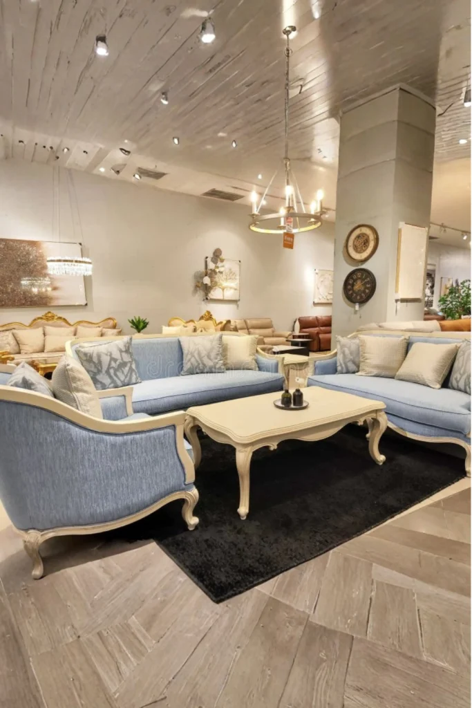 A furniture showroom display featuring a matching light blue living room set with a structured sofa, loveseat, and armchair arranged around a carved cream coffee table on a dark rug.