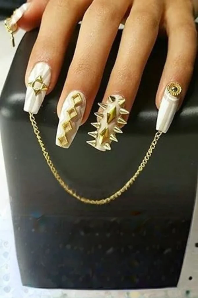 A close-up of a hand showcasing long, white square-shaped acrylic nails decorated with bold gold metal charms, spikes, and rivets. A delicate punk-style gold chain links the thumb nail to the pinky nail across the hand, highlighting edgy nail accessories design simple layouts to inspire your next creative manicure styling session.