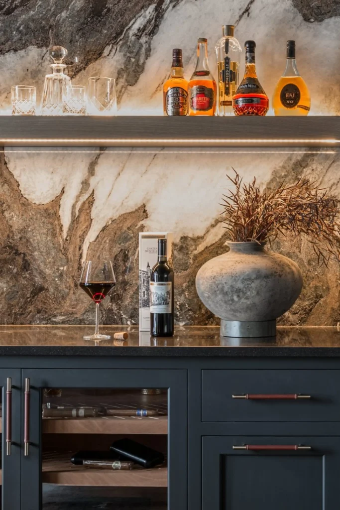 A moody home bar setup featuring a dramatic marble slab backsplash with rich metallic brown, bronze, and copper veining that creates a warm, textured look. Dark gray cabinets with leather-wrapped brass handles sit below a black countertop displaying a wine bottle, a filled glass, and an earthy stoneware vase with dried branches. An open shelf illuminated by glowing under-cabinet LED strip lights showcases liquor bottles and crystal decanters, capturing the essence of bold backsplash trends 2026.