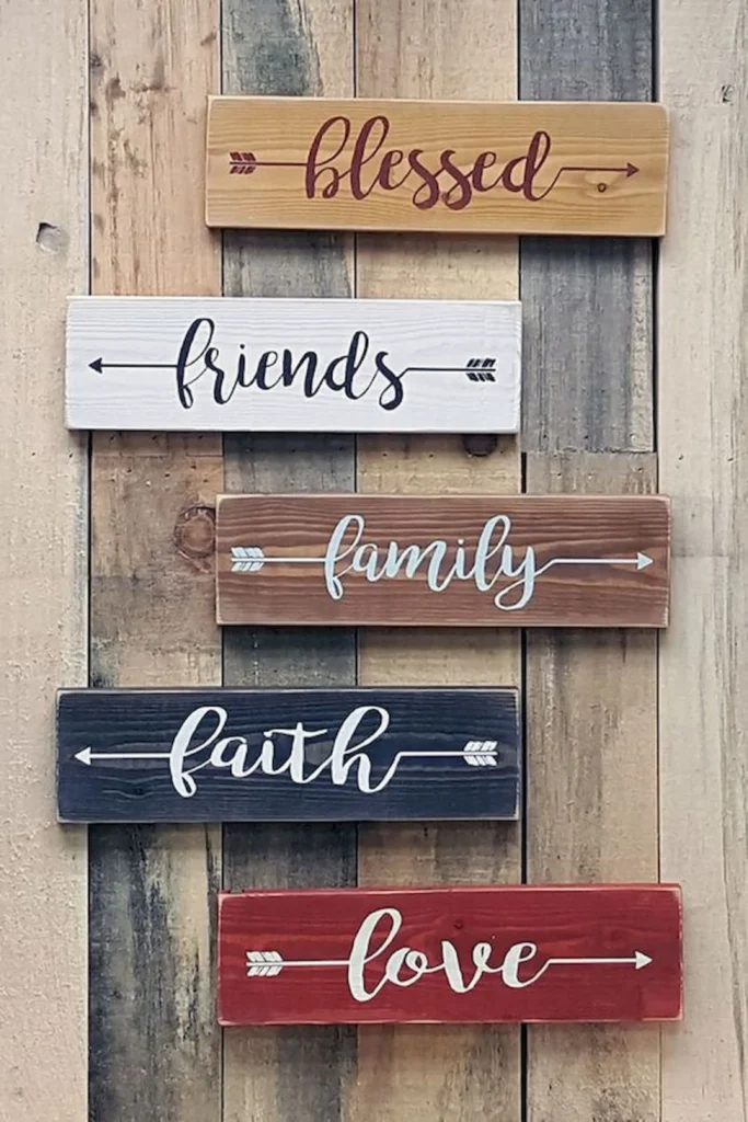 A vertical arrangement of five rustic wooden signs mounted on a wood plank wall, featuring cursive word art text including "blessed", "friends", "family", "faith", and "love" written across different colored blocks with decorative arrows.