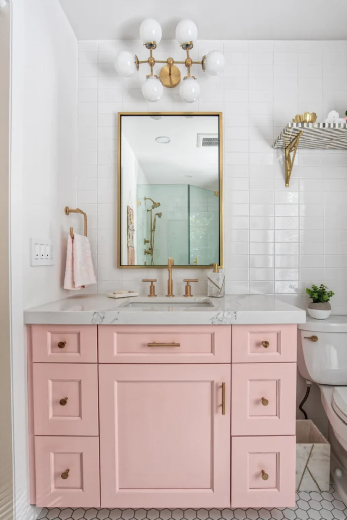 A chic, modern glam bathroom featuring a soft pink vanity topped with a luxurious slab of white marble with elegant gray veining. The pastel pink cabinet is adorned with brushed gold knobs and handles, matching the gold faucet, framed rectangular mirror, and the artistic sputnik-style light fixture on the white tiled wall above. A pink hand towel hangs on a gold ring to the side, showcasing how a blush or dusty pink vanity brings warmth and unexpected cheerfulness into an otherwise sterile bathroom, transforming it into a high-end, custom space.