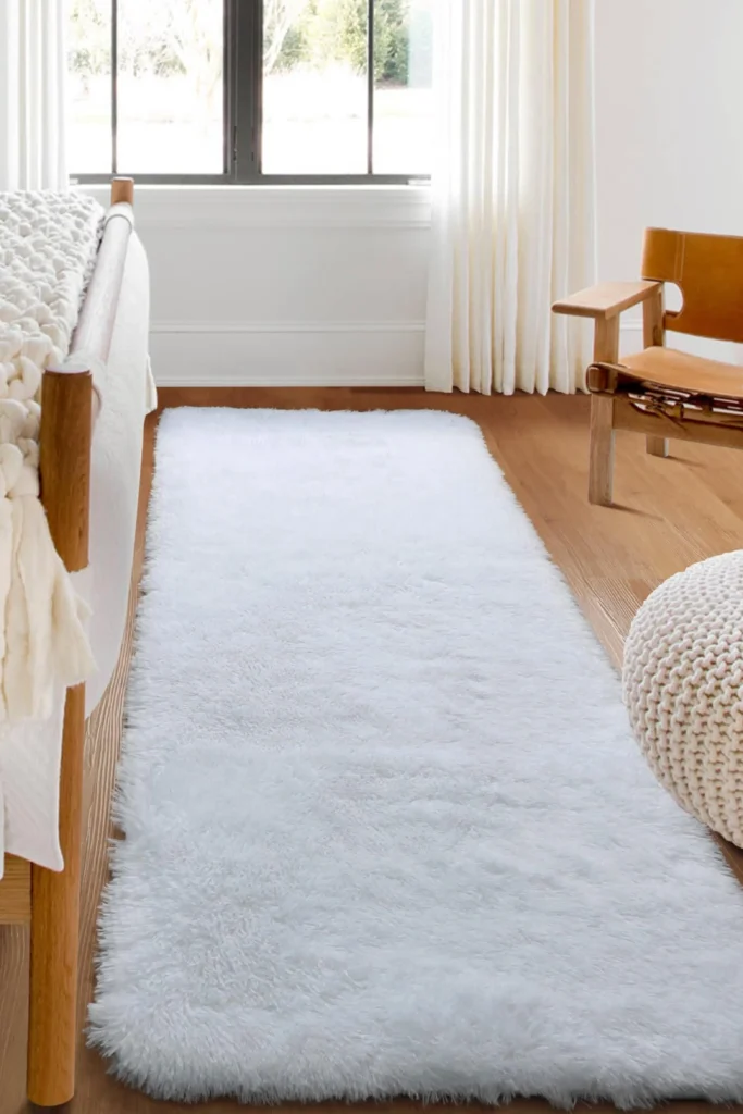 Plush Textured Rugs setup featuring a long, high-pile white faux fur runner rug placed over hardwood flooring right next to a wooden bed frame. The bedroom scene highlights a cozy approach to keep it soft with plush materials underfoot, complemented by a chunky knit blanket draped on the bed, a textured woven cream pouf in the foreground, and a minimalist wooden accent chair with a tan leather seat near a large window with cream curtains.