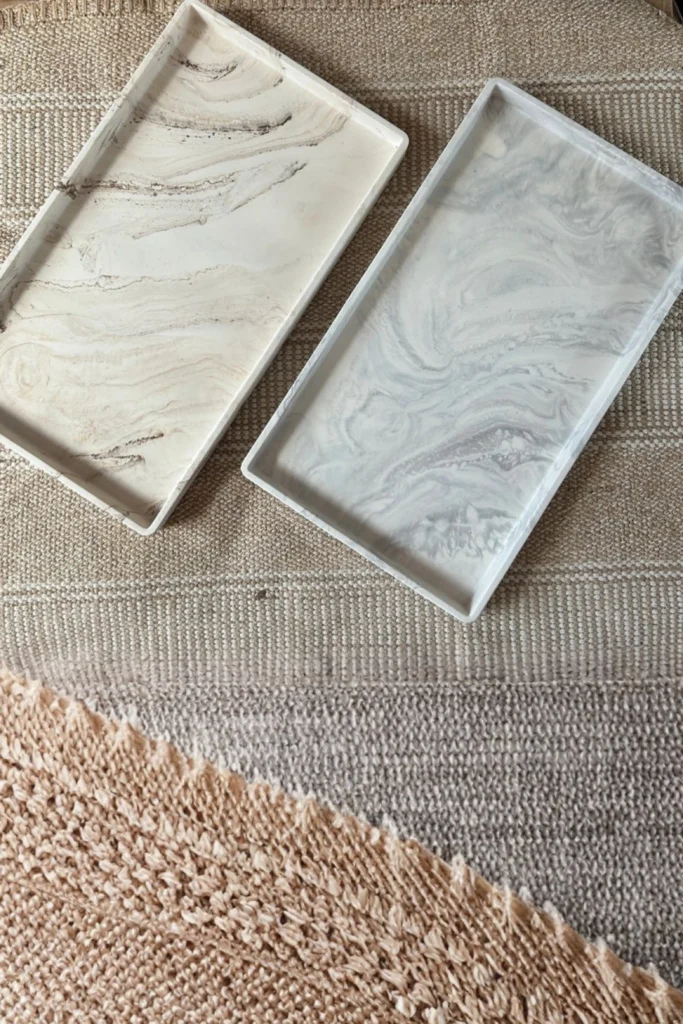 Two rectangular marble trays with organic swirling patterns displayed on a neutral woven textile background, showcasing that you do not need a solid marble countertop to enjoy a high-end feel. One tray features warm tan and beige marble veining, while the other features soft gray and white tones. Instead, place a heavy, rectangular marble slab or tray on your existing vanity. Use it as a beautiful base to display a high-quality scented candle and a small vase, instantly elevating your daily morning routine.