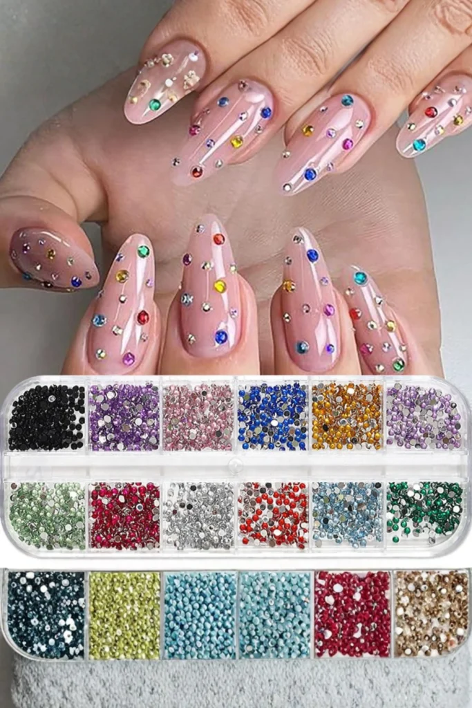 A close-up of long, nude almond-shaped nails decorated with multi-colored rhinestones. Below the hands, a clear plastic 24-compartment storage box is completely filled with sparkling multi-sized gems sorted beautifully by color and size, showcasing brilliant nail accessories ideas to organize your crystal collection and streamline your workspace.