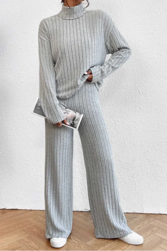 A woman wearing a gray turtleneck top and matching wide-leg pants from a collection of ribbed knit lounge sets, styled with white sneakers against a plain studio wall.