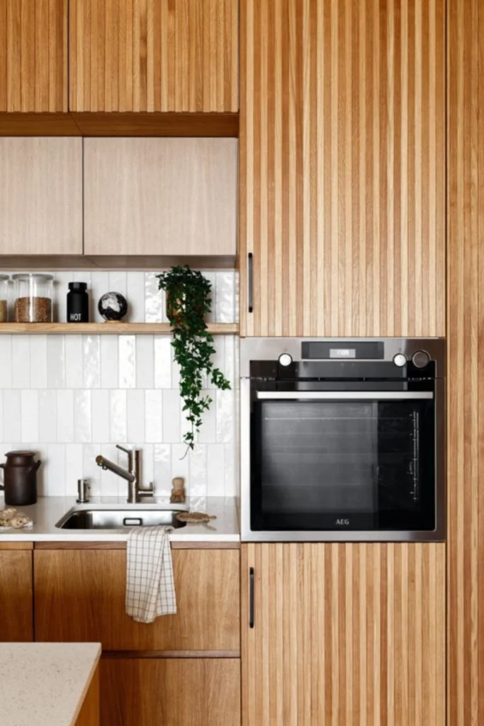 A modern kitchen showcasing ribbed and fluted textures with beautiful vertical ridges on light wood cabinets and a tall pantry surround. Next to an integrated stainless steel AEG oven, a small sink area features white vertical subway tile backsplash panels creating clean architectural lines. The space is styled with light countertops, open wooden shelving with kitchen decor, and cascading green plants, effortlessly guiding the eye upward to create a sense of linear depth and visual height.