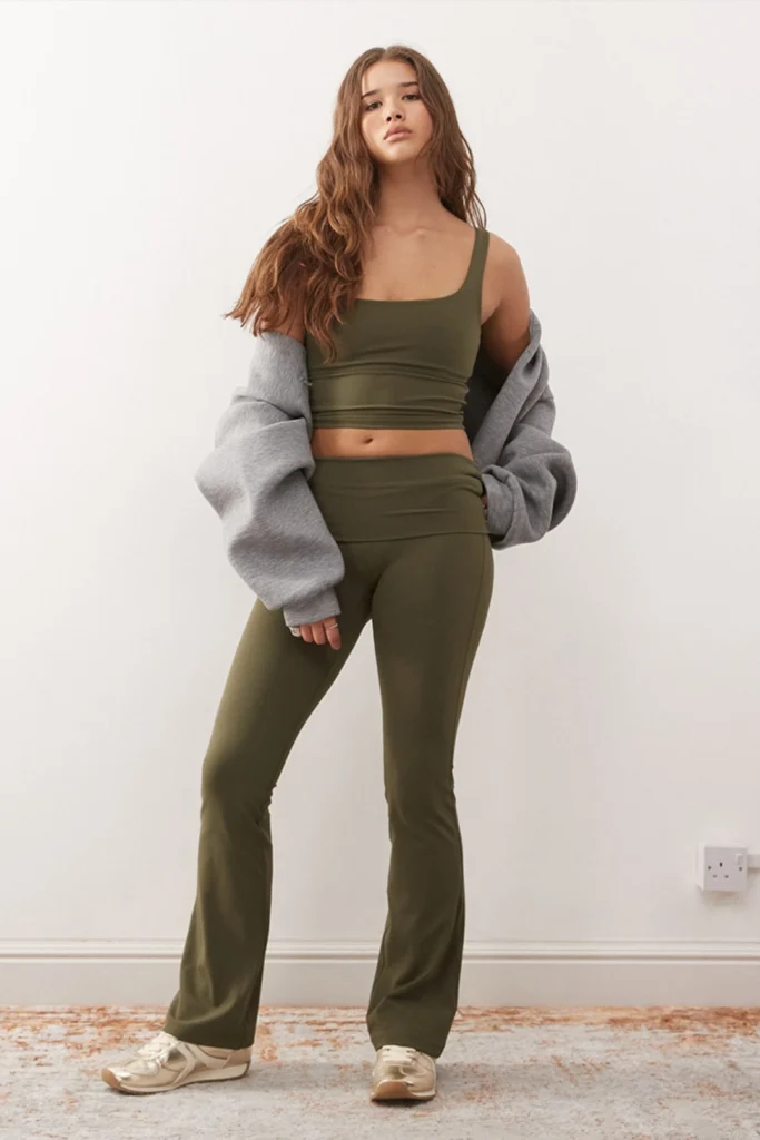 A woman posing in an olive green crop top and matching flared yoga pants from a collection of seamless activewear sets, styled with a gray hoodie and sneakers for comfortable home workout outfits women love.