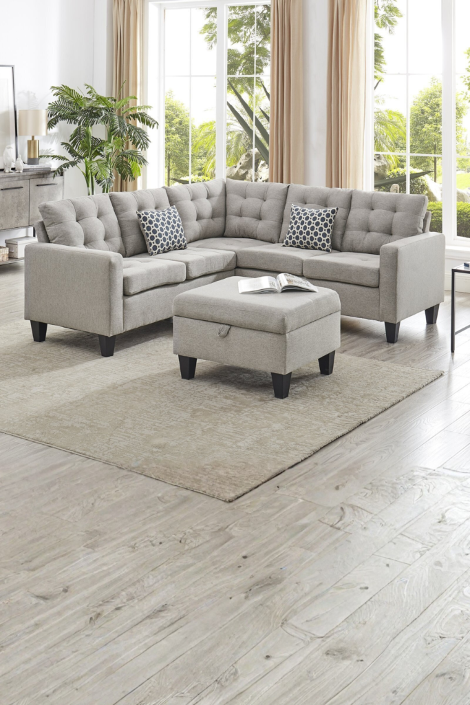 A modern living room layout featuring a light gray, tufted sectional sofa with matching patterned accent pillows and a central square fabric ottoman on a beige area rug. The furniture is beautifully arranged in front of large, bright floor-to-ceiling windows with soft beige drapes, showing creative furniture ideas for new home spaces.