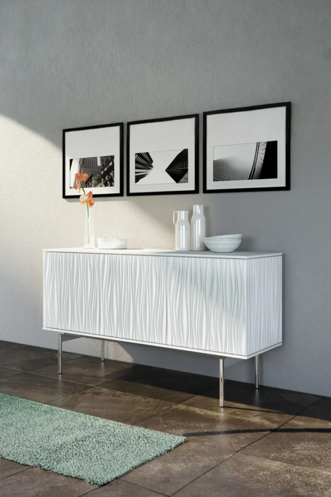 A modern white sideboard buffet cabinet with distinct textured, wavy cabinet doors and slim silver metal legs set against a gray wall. The tabletop is decorated with minimalist white ceramic pitchers, a bowl, and a slender flower vase, with a gallery wall of three square black-and-white architectural art prints hanging above, proving that keeping a brand-new space organized can be tough without the right storage solutions.