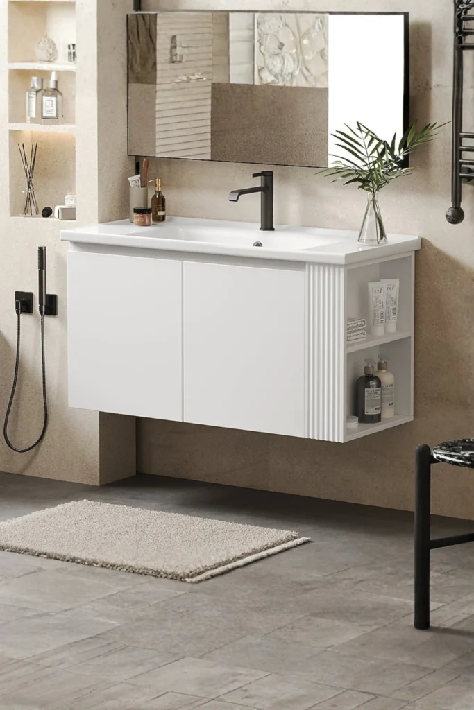 A modern minimalist bathroom showcasing a sleek white floating single vanity with an open bottom shelf on the side containing skincare bottles and folded towels. The vanity features a smooth white countertop sink with a matte black faucet, set against a textured beige wall. A large rectangular mirror hangs above the sink, reflecting light to create an open and airy feel, demonstrating gray small bathroom vanity ideas with mirror alternatives for a relaxed, spa-like aesthetic. A textured beige bath mat sits on the gray tiled floor.