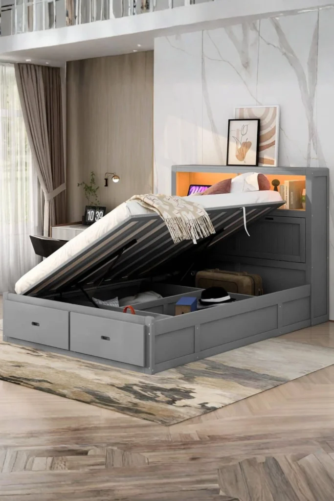 A gray wooden storage platform bed shown in a half-raised open position with hydraulic lifts, revealing built-in storage compartments under the mattress containing a suitcase and small boxes. The bed features an integrated headboard bookshelf with built-in ambient lighting, decor items, and a framed art piece, showcasing smart home organization and wonderful house warming gift ideas for new home furniture decoration.