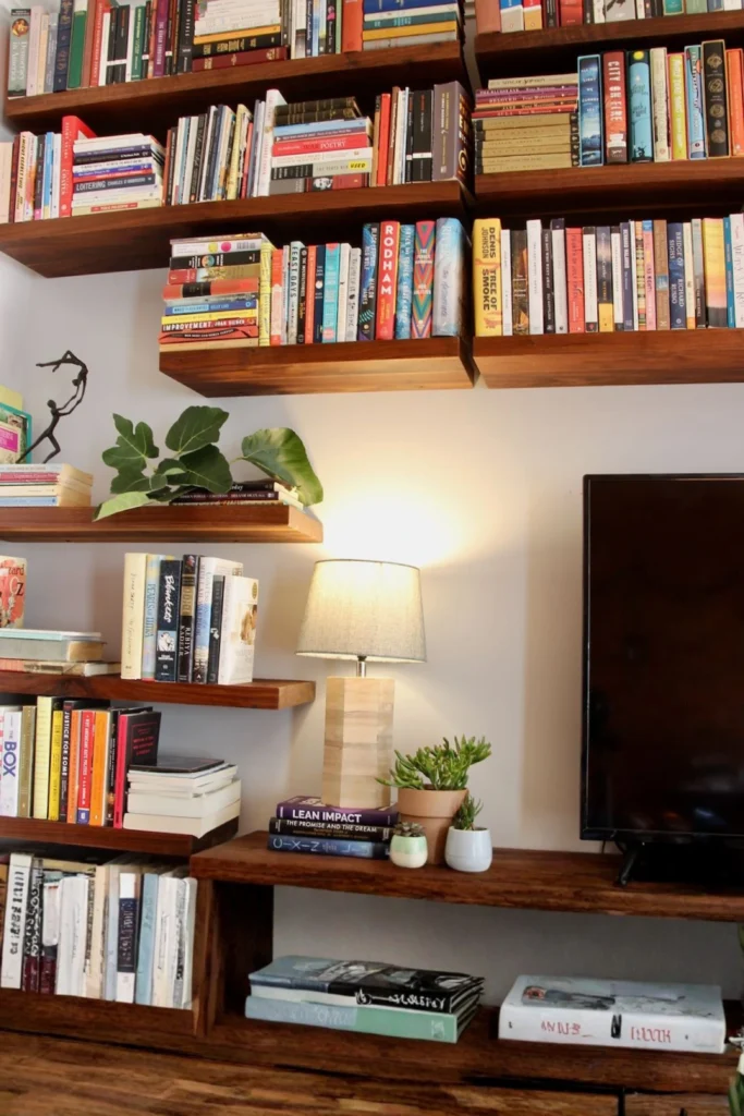A cozy home entertainment and library wall featuring staggered, dark wood floating shelves stacked vertically with a large collection of colorful books. Next to a flat-screen TV, a small table lamp with a gray fabric shade sits on a wooden console shelf, casting a warm glow over a few small potted green succulent plants. Display warm white books and cascading faux ivy vertically on floating wall shelves to draw the eyes upward. This clean, modern styling trick keeps your room feeling spacious yet deeply intimate during the dark winter months, as shown in Styled Floating Bookshelves