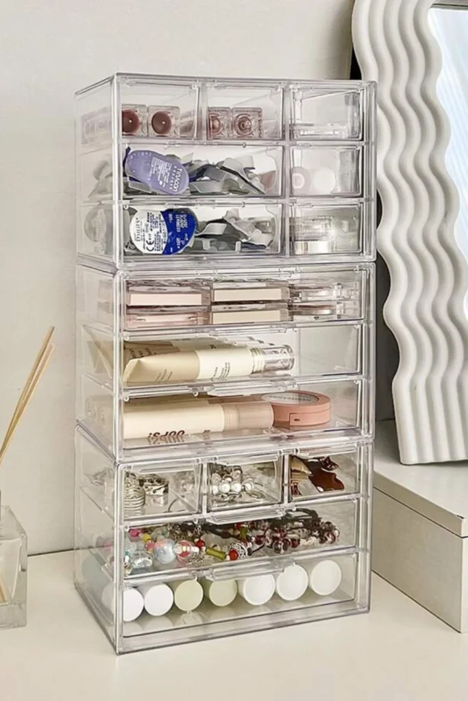 An organized vanity drawer setup showcasing clever beauty room ideas at home. The image features shallow, clear acrylic drawer dividers perfectly sorting makeup items. Eye palettes, compact powders, lipsticks, and foundations are neatly arranged lying flat and facing upward, creating a clean, satisfying, and easily accessible display inside a white drawer.