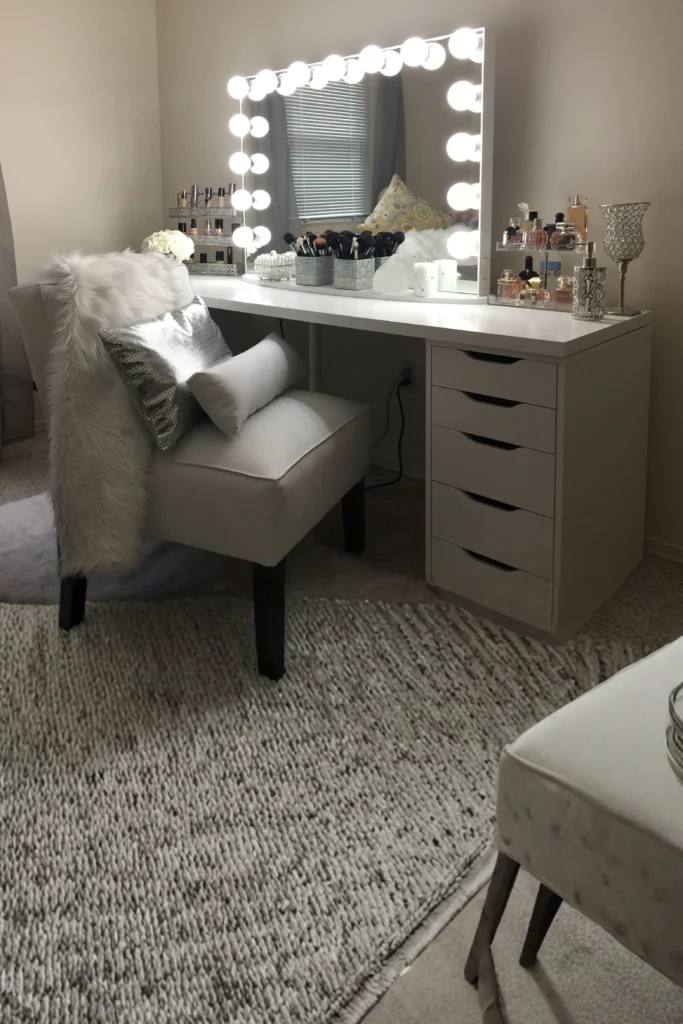 A beautiful makeup vanity station highlighting modern beauty room ideas at home. The setup features a large rectangular tabletop Hollywood mirror surrounded by glowing, frosted white LED bulbs that provide even lighting. The white desk is organized with acrylic makeup trays, perfume bottles, and brushes, creating a professional studio feel next to a bright room window.