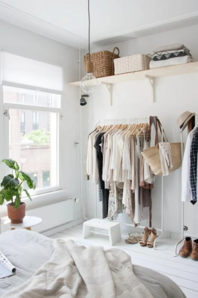 A bright, airy boutique-style room from the image "The Modern Capsule Display Setup.webp" highlighting a curated minimalist clothing style women adore. The space features a freestanding white metal garment rack holding neutral, cream, and black clothing on uniform wooden hangers alongside a hanging woven straw tote bag. Above the rack, a light wood floating shelf holds storage baskets and folded knits, while a large window lets in natural light onto white wooden floors, a small potted green plant, and a neatly made bed in the foreground.