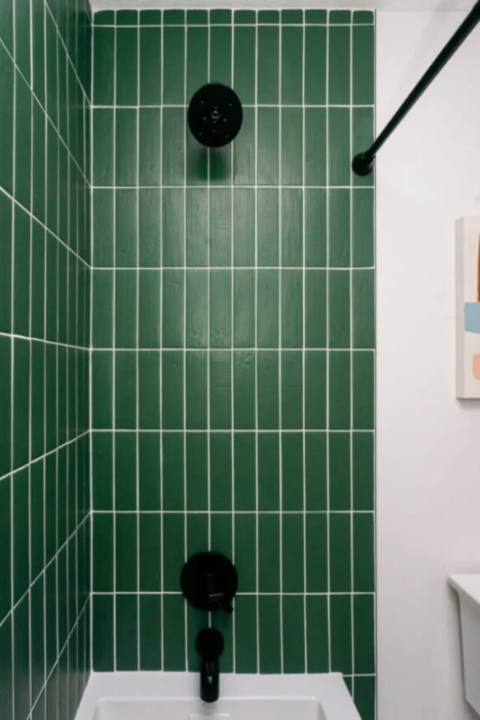 A contemporary bathroom corner showcasing a vertical stacked tile layout using slim, forest green rectangular subway tiles with crisp white contrasting grout. This architectural style, reflecting modern backsplash trends kitchen and bath aesthetics, draws the eye upward to make the ceiling look taller. The vibrant green tile grid is paired with minimalist matte black fixtures, including a round shower head, a control knob, a tub spout, and a black shower rod against a clean white wall.