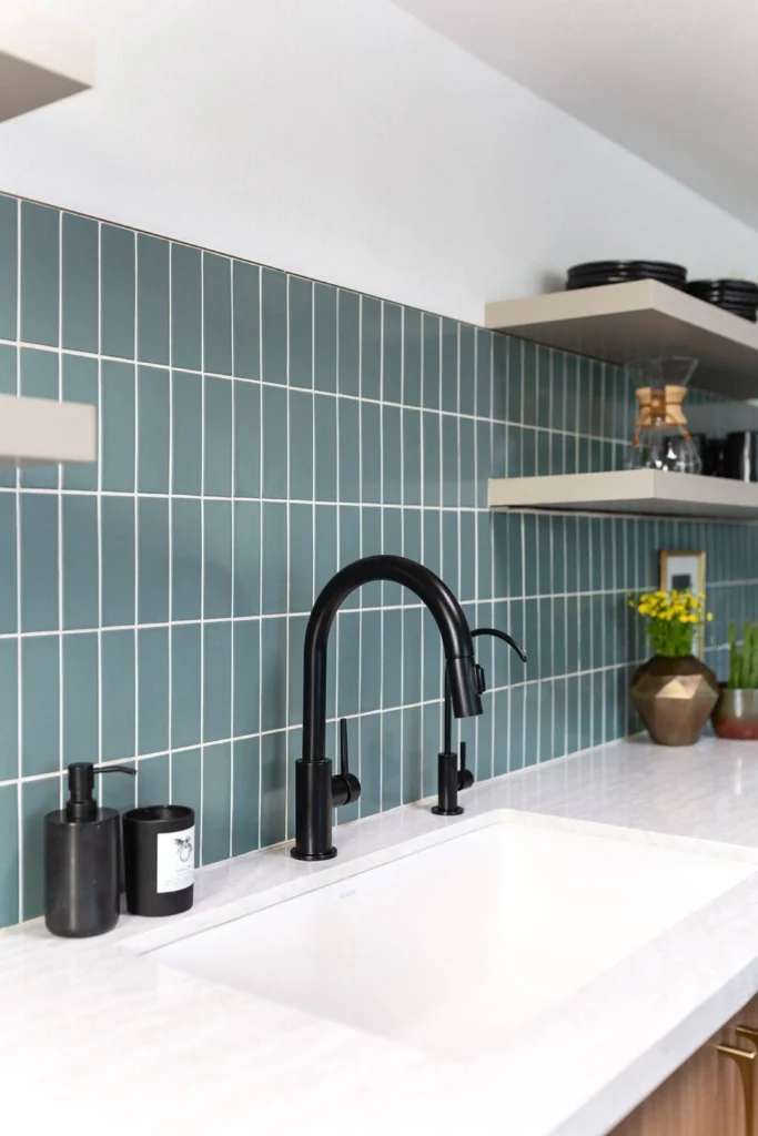 A contemporary kitchen space featuring a vertical subway tile stacks backsplash in a muted teal or slate blue color. The elongated rectangular tiles are stacked vertically side-by-side with contrasting white grout lines, creating crisp, clean parallel lines that show a modern alignment. The setup includes a white undermount sink, light marble countertops, a sleek matte black gooseneck faucet with matching soap dispensers, and open floating shelving holding minimal kitchen decor.