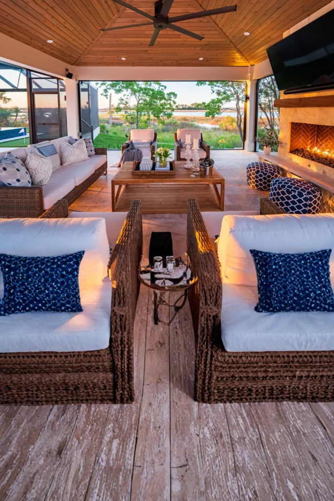 A covered luxury patio with a wooden vaulted ceiling and fireplace, featuring wicker sofas and armchairs with white cushions and blue patterned pillows oriented toward a scenic view of a green landscape and water.