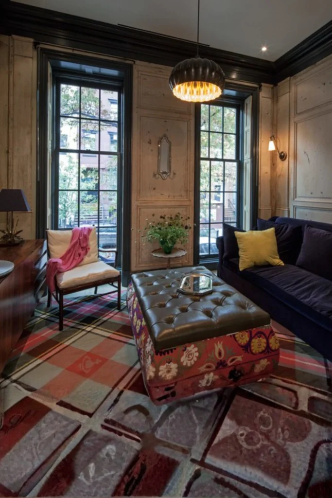 A moody, eclectic living room interior featuring an expansive plaid patterned carpet floor matching the aesthetic of Warm Plaid and Tartan.jpg. The room is styled with a dark navy blue velvet sofa adorned with a single mustard yellow throw cushion, facing a tufted leather ottoman coffee table wrapped in an ornamental floral fabric base. In the background, tall black-framed multi-pane windows look out to a street view, set against rustic wood-paneled walls with dark wood trim elements and elegant ambient lighting. Add a subtle plaid patterned accent blanket or a couple of tartan cushions to your main seating area. This classic pattern evokes immediate lodge-style nostalgia and pairs beautifully with dark wood elements.