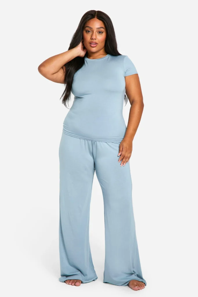 A plus-size woman with long dark hair posing barefoot against a solid white background. She is wearing a soft, dusty blue loungewear set featuring a crew-neck short-sleeved top and loose, flowing wide-leg modal trousers that offer a comfortable and luxurious high-end feel.
