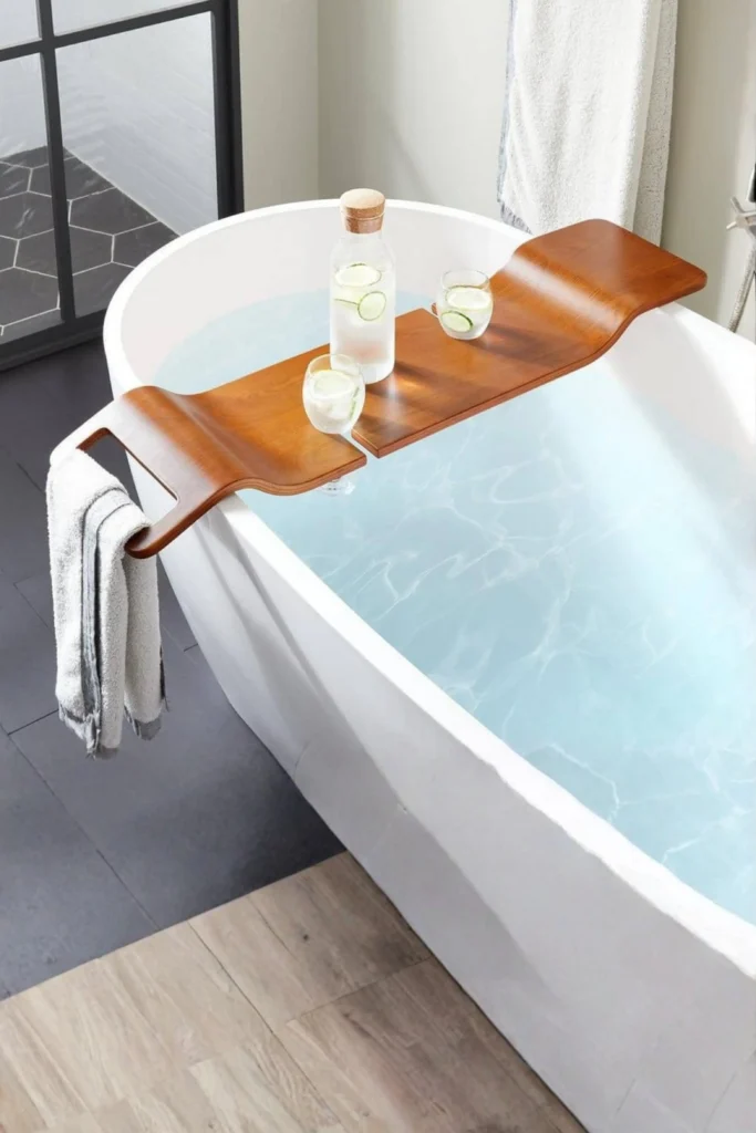 A sleek, curved warm wooden bath bridge resting across a modern freestanding white bathtub filled with clear water, highlighting how to balance the cool, smooth texture of stone, pair your marble pieces with a warm wooden bath bridge or caddy over the tub. A grey towel hangs from the built-in handle side, while the tray holds a glass carafe and two matching glasses filled with cucumber-infused water. This combination of materials brings a rich, layered texture to the room. It provides the perfect placeholder for a book or a glass of water during a relaxing soak.