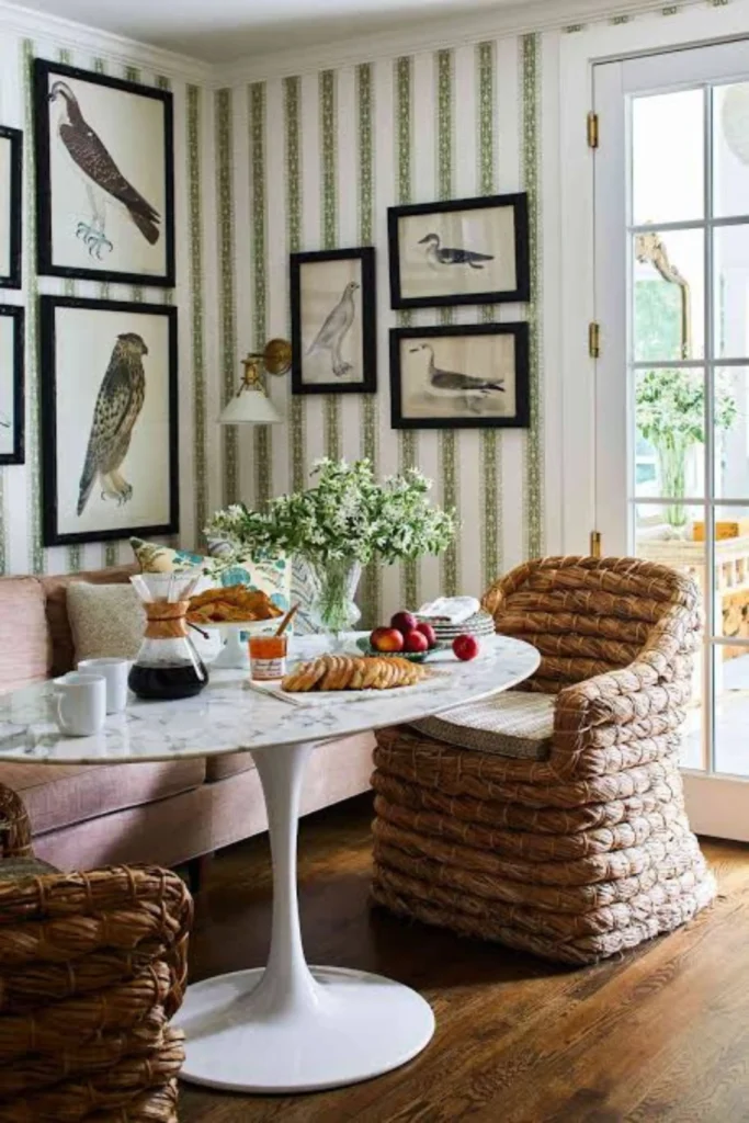 An elegant dining space highlighting a striking Accent Wall Breakfast Nook concept. The design features a white marble oval tulip table paired with chunky woven seagrass armchairs and a mauve pink upholstered banquette sofa. The back wall serves as a focal point with green and white striped wallpaper decorated with a gallery layout of vintage framed bird art prints.