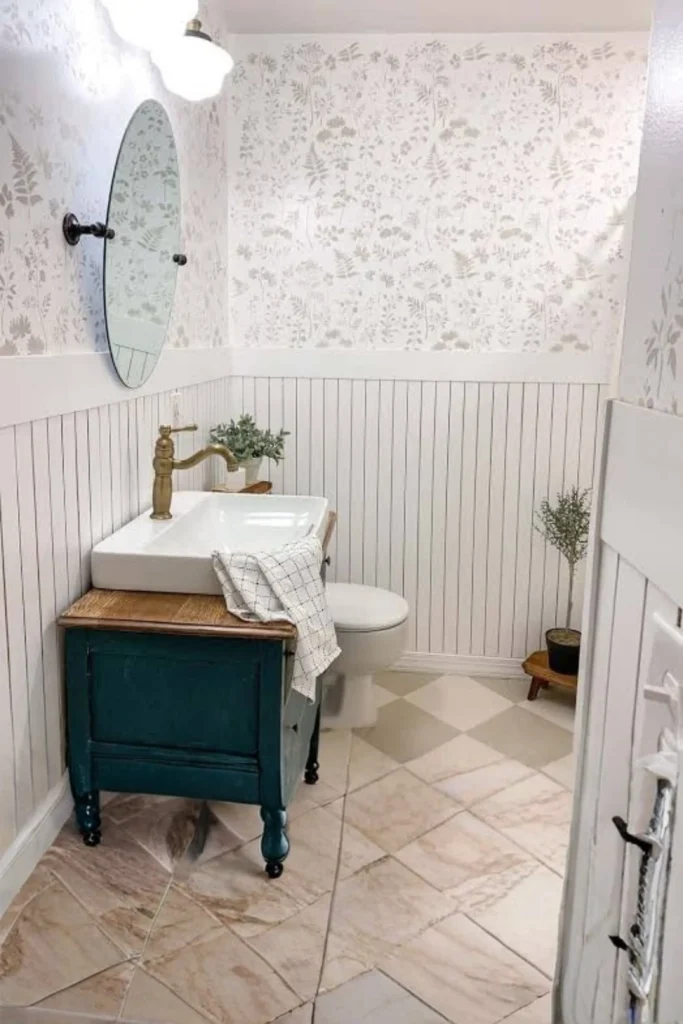 A cozy farmhouse powder room showcasing gray bathroom remodel ideas 2026 trends with vertical white beadboard wall paneling. The beadboard wainscoting runs halfway up the wall, capped with a white wood ledge that meets a soft gray floral patterned wallpaper. An antique dark teal vanity with a brass faucet and white vessel sink rests on neutral diamond-patterned floor tiles under an oval tilt mirror.