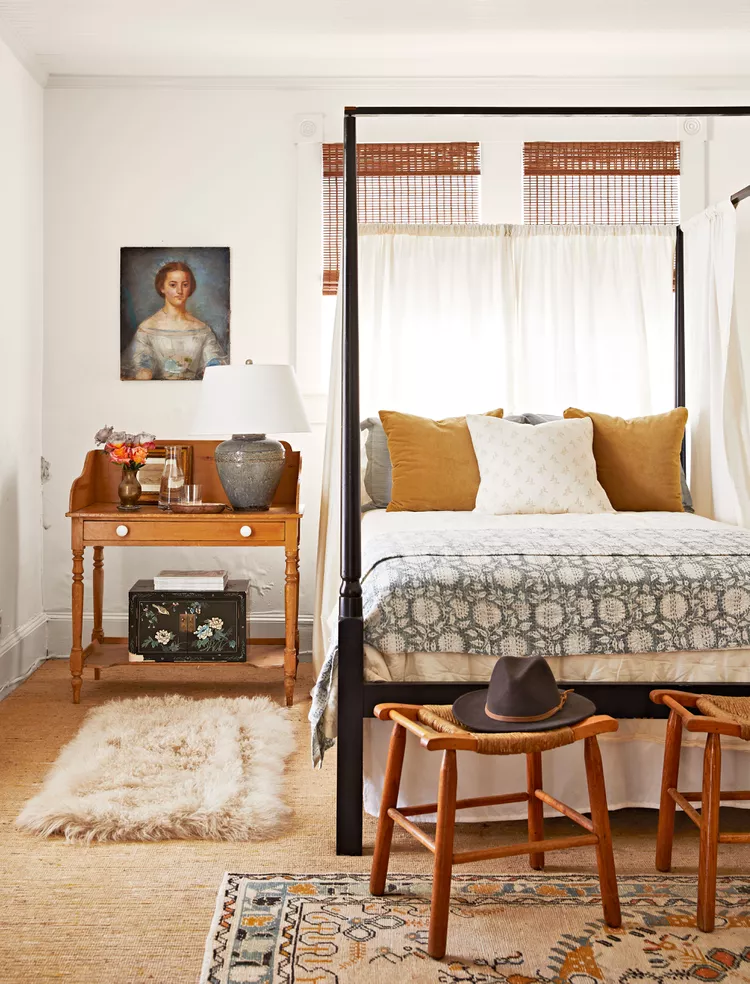 A cozy vintage bedroom layout showcasing clever bedroom flooring ideas by choosing to add layers over a hard floor. The space features a base layer of a textured natural flat-weave rug beneath a black four-poster canopy bed. A smaller, plush white faux-fur shag runner rug is layered as an accent texture along the side of the bed next to an antique wooden vanity desk, while a patterned bohemian area rug anchors the foreground layout.