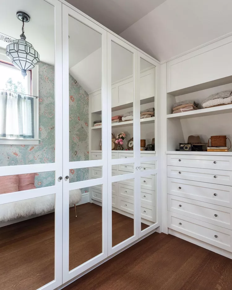 A bright, custom walk-in dressing room inside a sloped room, featuring medium-toned wood floors and white cabinetry. Large floor-to-ceiling wardrobe doors fitted with custom-cut mirrored panels perfectly reflect a patterned wallpapered wall, a window, and open storage shelves across from them, demonstrating creative attic closet ideas low angled ceilings to bounce light and make a narrow layout feel twice as wide.