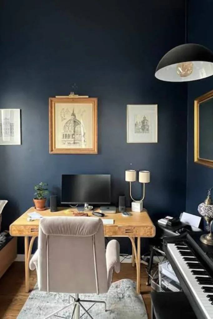 A sophisticated workspace illustrating navy blue accent wall ideas to create a professional background. The setup features a rich oak writing desk positioned directly in front of a deep navy blue accent wall in home office ideas layout. A comfortable light grey office chair sits on a distressed area rug, while framed artwork, a small potted plant, a unique U-shaped desk lamp, and a large black dome pendant light complete the high-end executive look.