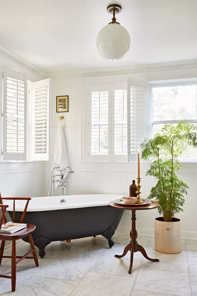 A bright, classic bathroom setup showing how to add shutters on windows for architectural depth and privacy. The scene features white plantation window shutters framing a corner with a vintage black clawfoot soaking tub. The room is decorated with white shiplap wainscoting, a light marble tile floor, a round wooden side table with a lit candle, and a large green potted plant under a modern frosted globe pendant light.