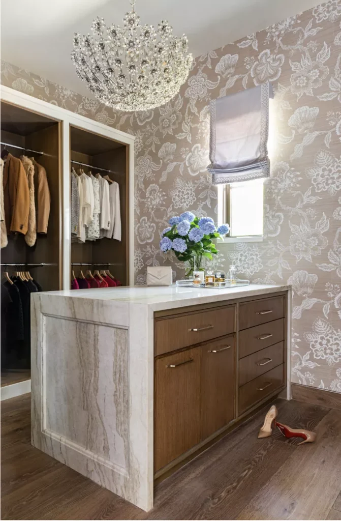 A luxury boutique-style walk-in dressing room featuring a large central island with wooden drawers and a thick, white marble waterfall countertop. The room is decorated with a stunning crystal floral chandelier and a textured, dusty rose wallpaper accented with large white damask floral patterns, showing how adding a beautiful, stylish wallpaper turns a basic storage area into a luxury sanctuary.