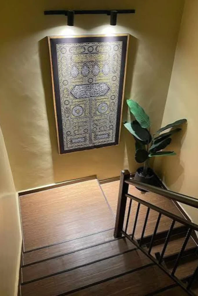 A high-angle view of a modern stairwell showcasing staircase landing ideas to brighten dark corners. The landing features smooth warm-toned walls and a large, vertically hanging black and gold Islamic calligraphy art piece illuminated by sleek black overhead track spotlights. In the corner of the wood-textured landing platform, a large potted green faux plant adds vibrant life next to a dark wooden stair railing.