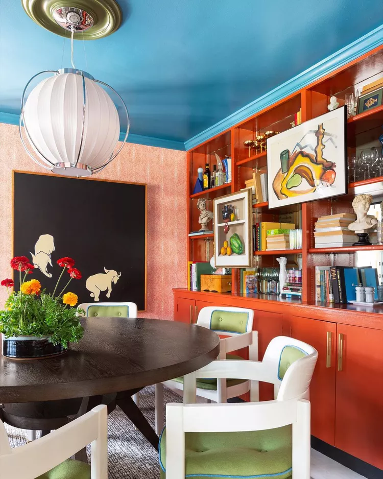 A vibrant, colorful dining room with an orange wooden hutch, pink patterned wallpaper, and a round wooden dining table, styled with a bold contrasting painted ceiling in a striking bright turquoise blue that extends seamlessly down to the crown molding to show how a bold color choices can add instant moodiness and high-impact style on a budget.