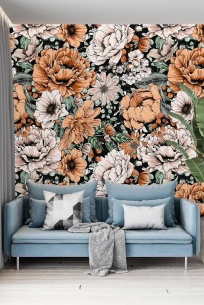 A striking living room display illustrating the concept of Botanical Maximalism and Oversized Florals. The setup features a bold accent wall covered in a dramatic, dark wallpaper packed with large-scale peach, cream, and orange peony flowers. A modern light-blue sofa with styled throw pillows sits directly in front of the vibrant floral pattern wall, creating a custom sanctuary vibe.