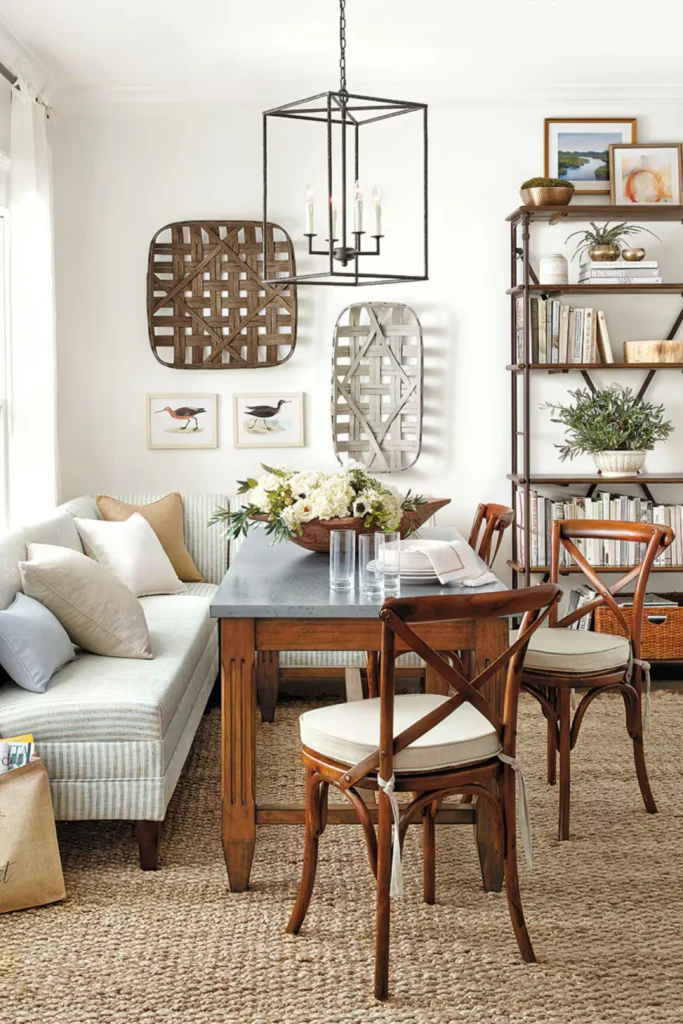 A rustic farmhouse dining space showcasing a cozy Breakfast Nook Banquette layout. The design features a long rectangular wooden table with a grey top, paired with classic X-back wooden chairs and a light blue striped upholstered banquette sofa bench loaded with throw pillows. The white walls are decorated with woven tobacco baskets and small bird prints beneath a large black iron lantern pendant light.