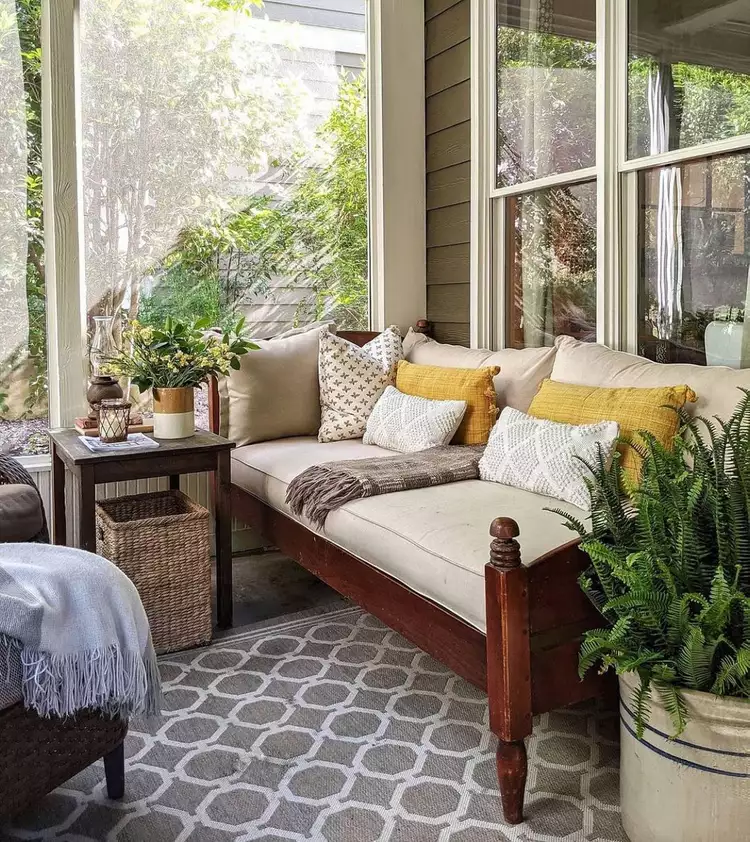 A relaxing resort-style setup for small screened-in porch ideas cozy nooks, featuring a vintage wooden twin daybed styled with mustard yellow and patterned throw pillows, a grey geometric rug, and a large potted fern.