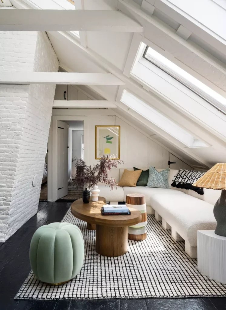 A bright, modern attic living room featuring a sloped ceiling with integrated large glass skylights and white-painted exposed beams. Natural light floods a cozy white sectional sofa, custom wooden coffee tables, and a pastel green pumpkin-shaped ottoman on a patterned rug, illustrating how beautiful false ceiling ideas with skylights and beams bring the beauty of the outdoors inside.