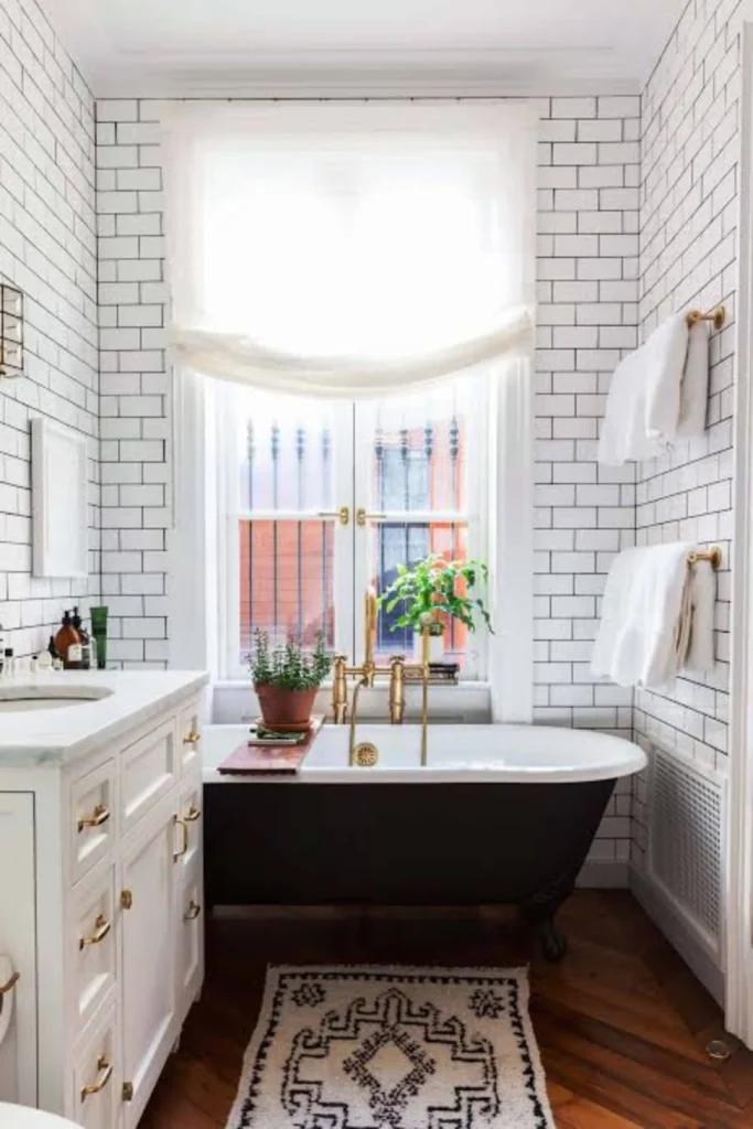 A bright industrial-style bathroom layout highlighting smart bathroom remodel ideas on a budget. The walls are covered from floor to ceiling in classic white subway tiles with a bold, contrasting deep charcoal grout. A luxury black clawfoot bathtub with elegant brass fixtures sits underneath a large window, next to a white vanity cabinet with marble countertops and brass drawer handles on rich hardwood floors.
