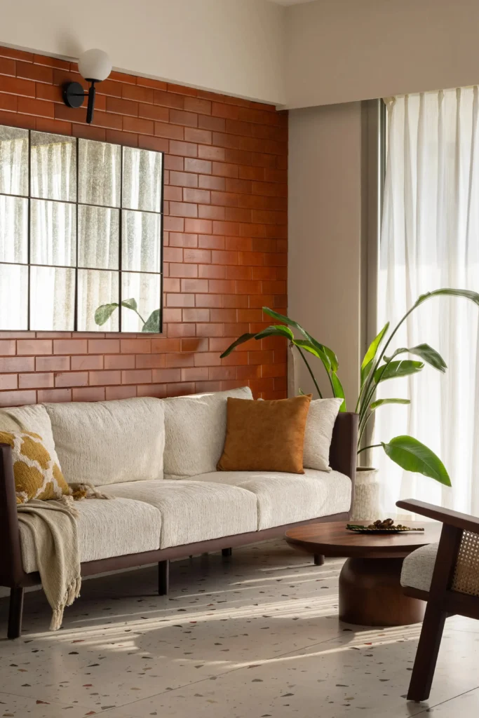 A bright mid-century modern living room showcasing modern house decorating ideas with a cream-colored textured sofa. The couch is complemented with an earthy brown throw pillow, a yellow patterned cushion, and a soft knit throw blanket. The background features a striking terracotta brick accent wall with a grid mirror, a wooden coffee table, a large indoor plant, and elegant terrazzo flooring.