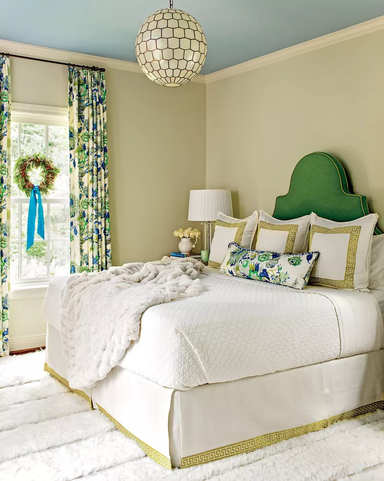 A beautifully styled bedroom illustrating how to use soft bedroom flooring ideas by choosing tactile textures to prevent a neutral space from looking boring. The room features a dense, ultra-thick white ribbed textured carpet that adds depth under a white bed with a bright green headboard. The layout is completed with a plush white faux-fur blanket throw, light beige walls, a blue-painted ceiling, and an architectural geometric honeycomb globe pendant light hanging above.