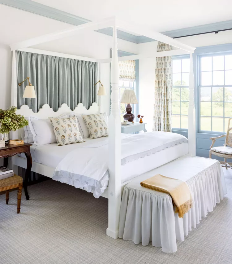 A serene blue and white master bedroom layout demonstrating how to choose practical bedroom flooring ideas by considering your lifestyle. The room features a low-profile light gray plaid patterned low-pile carpet flooring that provides a soft foundation under an elegant white four-poster canopy bed. Light enters through blue-trimmed double windows decorated with floral curtains, beautifully matching a soft gray headboard curtain drape.