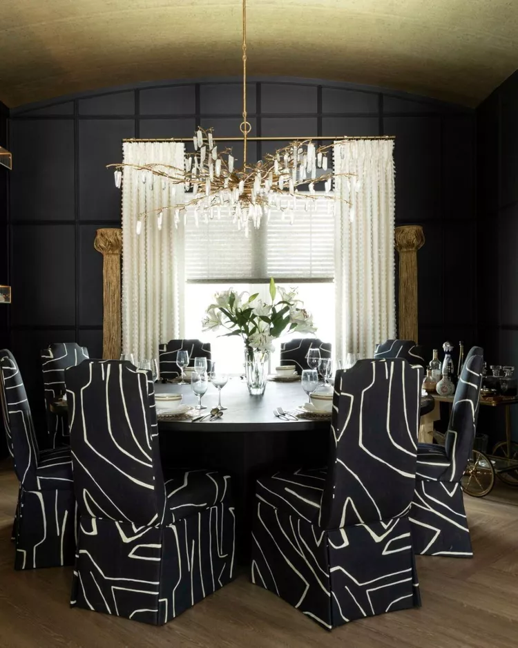 A moody, sophisticated dining room featuring black paneled walls, a round dining table with abstract black-and-white patterned slipcovered chairs, and a striking gold branch crystal chandelier, all crowned by a curved ceiling lined with textured cork-style wallpaper to show how this organic material brings a warm, earthy bohemian aesthetic to a high-end design.