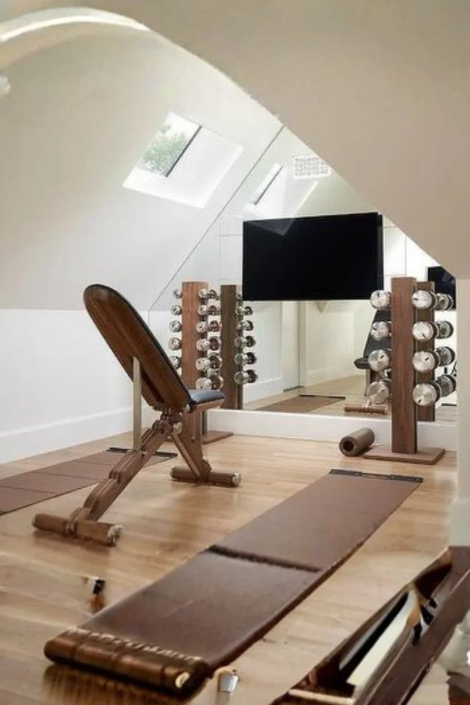 An attic home gym with angled walls featuring a custom corner mirror setup that reflects a wooden workout bench, vertical dumbbell racks, a mounted TV screen, and light pouring in from a ceiling skylight.