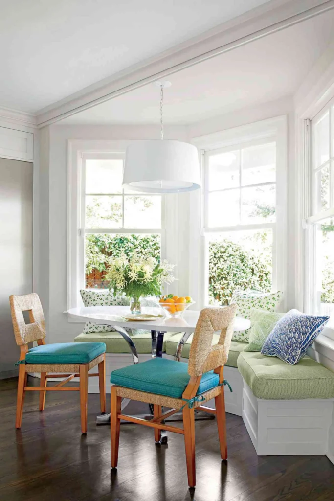 A bright kitchen bay corner displaying a vibrant Corner Breakfast Nook setup. The design features a white oval tulip-style table with chrome legs, paired with two woven rattan chairs hosting turquoise cushions. An L-shaped white built-in storage banquette bench fits neatly into the window corner, decorated with light green cushions, patterned throw pillows, and a clean white drum pendant light overhead.