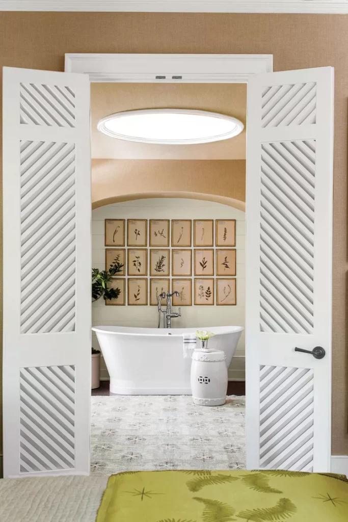 A serene master bath layout viewed through open white louvered doors, demonstrating how to create your own view using wall decor. A modern white freestanding soaking tub sits below an arched alcove wall decorated with a large grid of framed botanical pressed-leaf prints. The room features warm tan walls, a round white ceramic garden stool, a soft patterned area rug, and an architectural round skylight illuminating the layout.