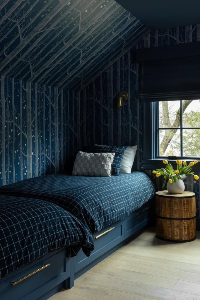 A cozy attic bedroom showcasing moody navy blue accent wall ideas with forest-patterned wallpaper wrapping around a sloped ceiling. The space features built-in twin beds with matching navy blue gridded comforters and storage drawers, a warm brass wall sconce, and a round wooden side table holding a vase of bright yellow tulips next to a window.
