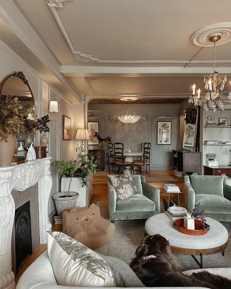 An elegant, classically styled living room with soft sage-green velvet armchairs and an ornate fireplace mantel, showcasing intricate DIY ornate molding on the ceiling with delicate corner appliques and a matching plaster ceiling rose surrounding a crystal chandelier to bring old-world European charm to life.